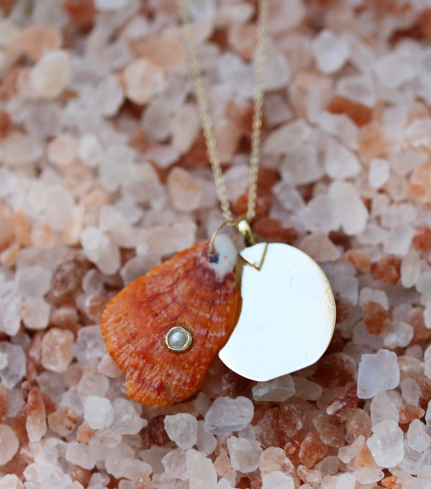 Collier en or avec un coquillage orange et une breloque lune blanche, sur des cristaux de sel roses.