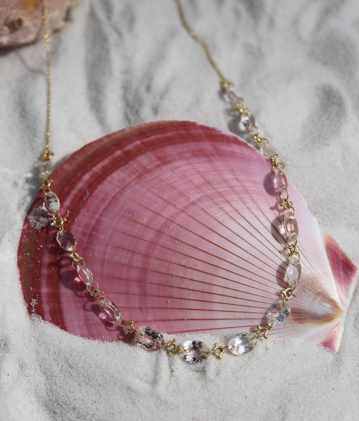 Collier doré avec pierres précieuses ovales transparentes drapées sur un coquillage rose dans le sable.