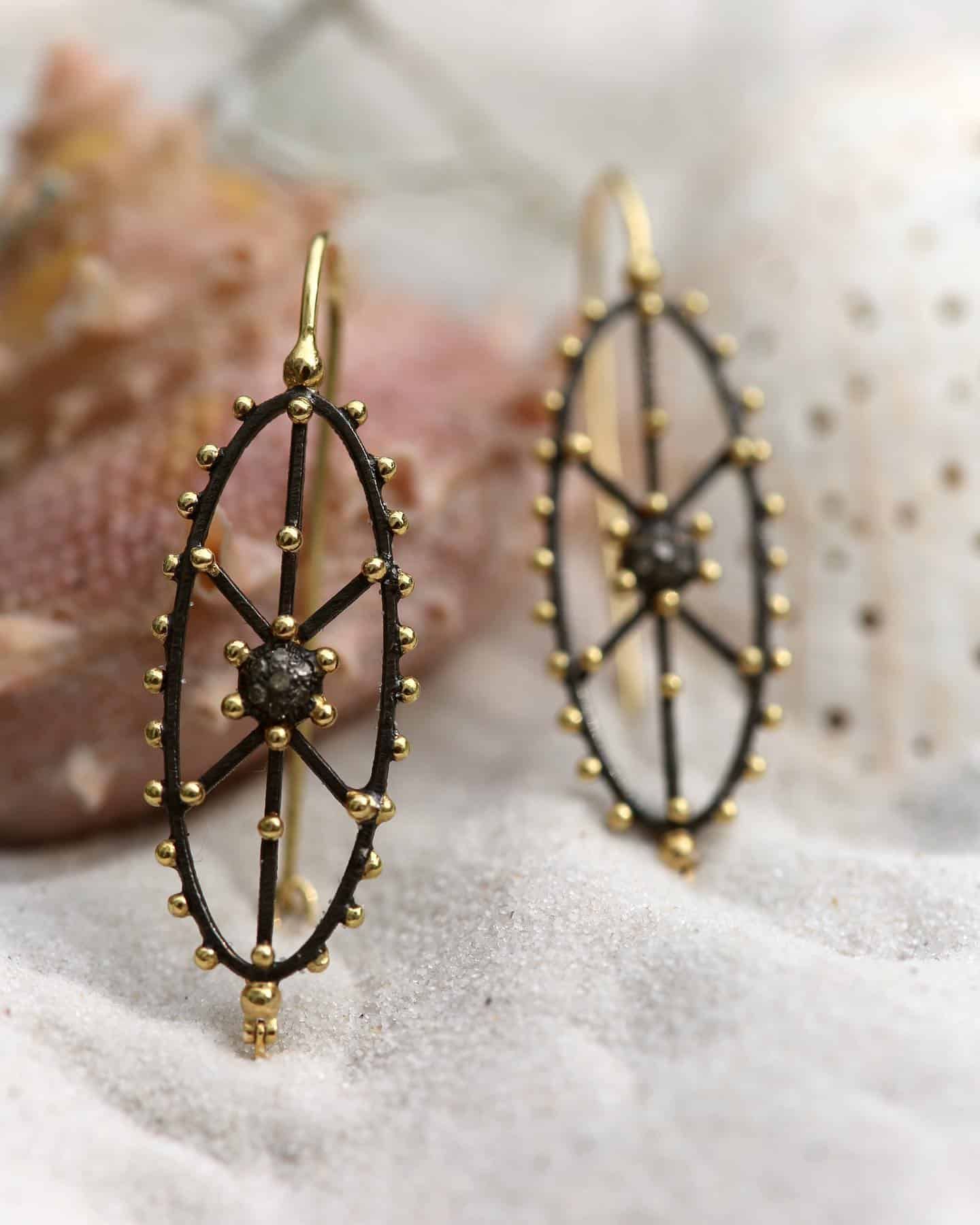 Boucles d'oreilles en filigrane doré et noir avec un motif en étoile, sur un lit de sable blanc.