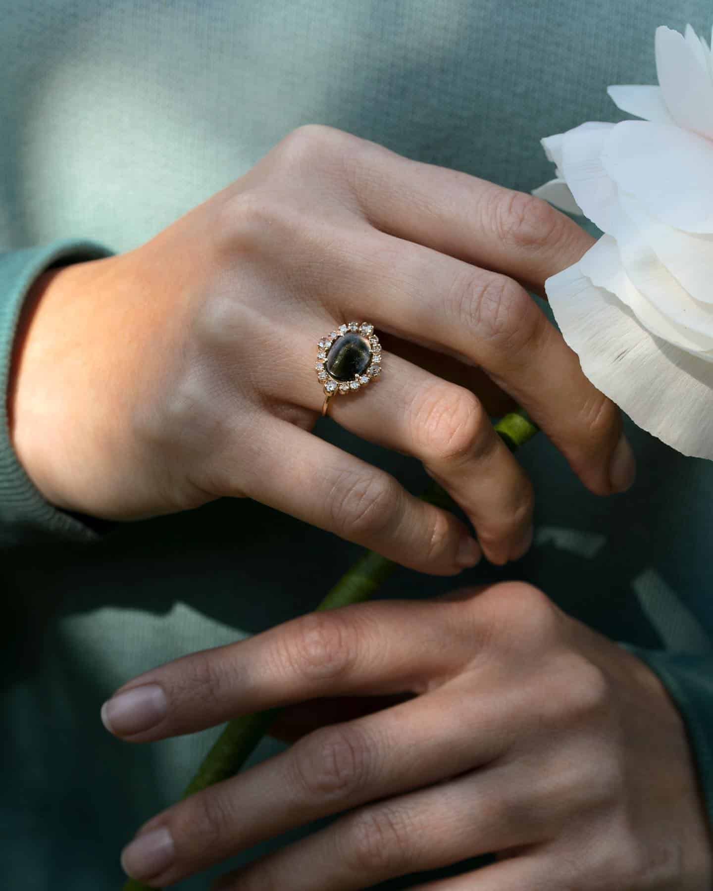 Main avec une bague en pierre sombre tenant une fleur blanche sur un fond bleu clair.