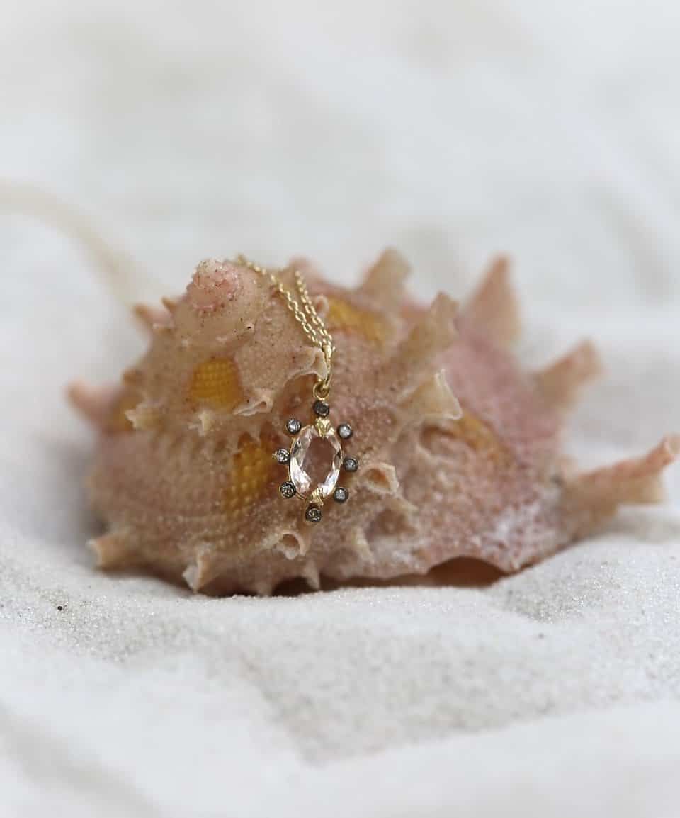 Collier en or avec un pendentif rose sur un gros coquillage beige, posé sur du sable blanc.