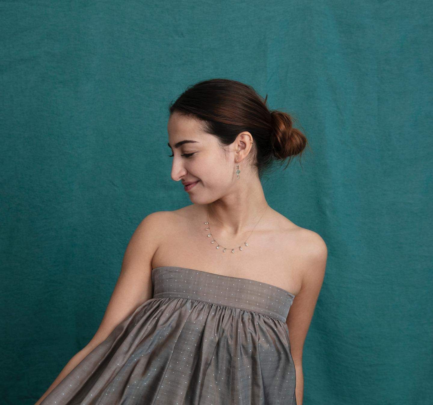 Une femme vêtue d'une robe grise sans bretelles sourit, regardant vers le bas, les cheveux en chignon sur un fond bleu sarcelle.