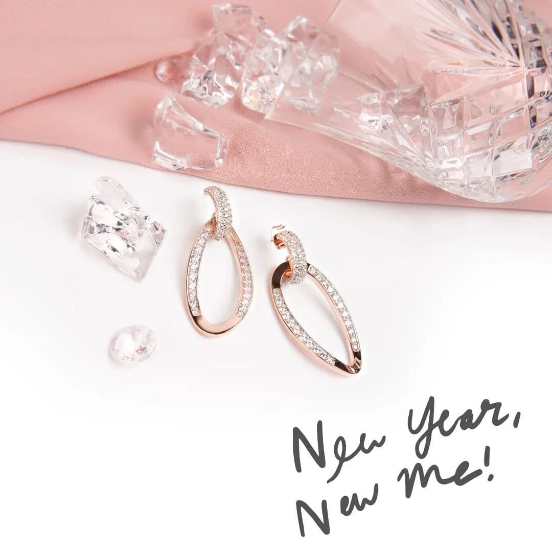 Paire de boucles d'oreilles en or rose avec pierres scintillantes, disposées sur fond blanc avec tissu rose et glace.