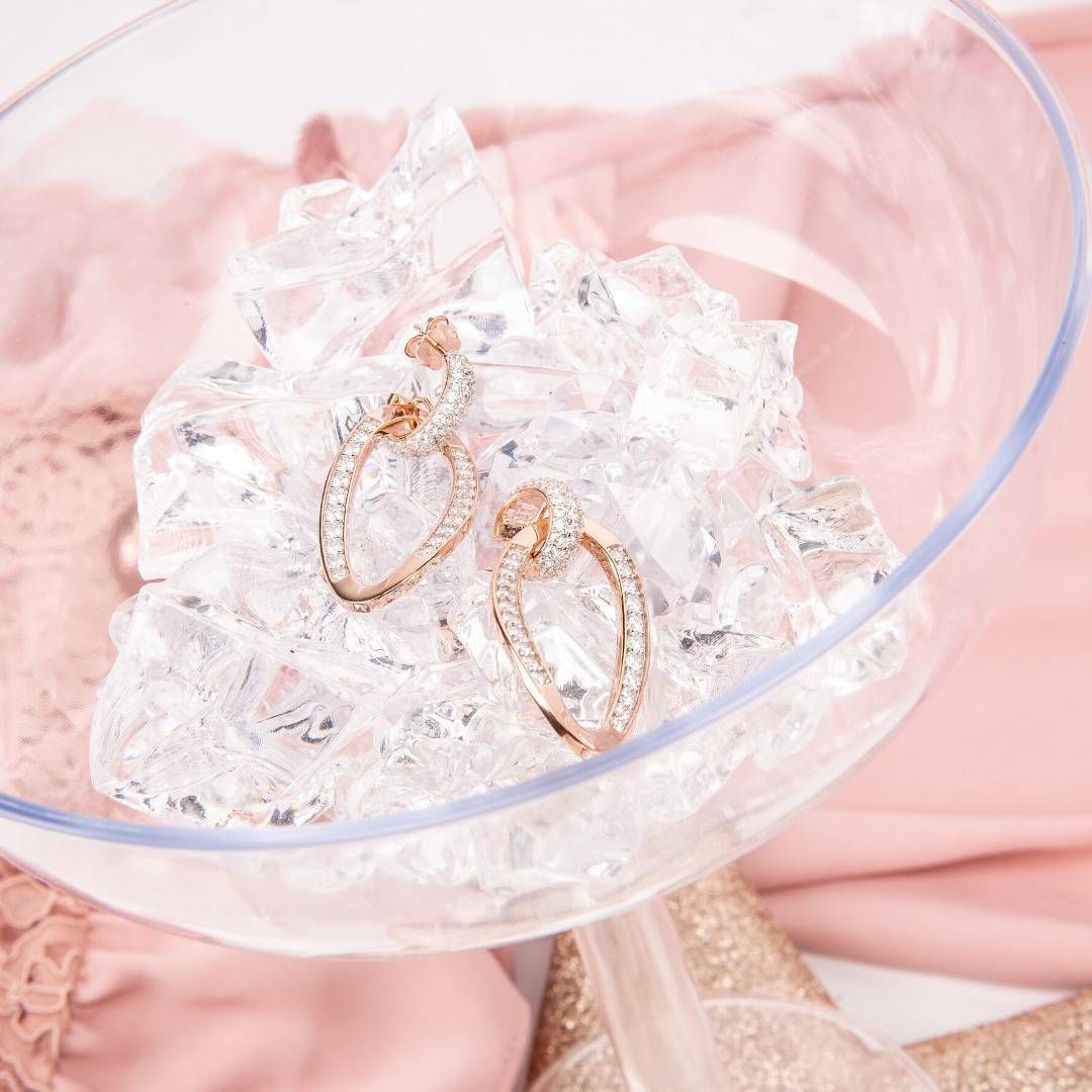 Boucles d'oreilles créoles en or rose avec pierres précieuses transparentes dans un verre à champagne rempli de glace pilée, sur tissu rose.
