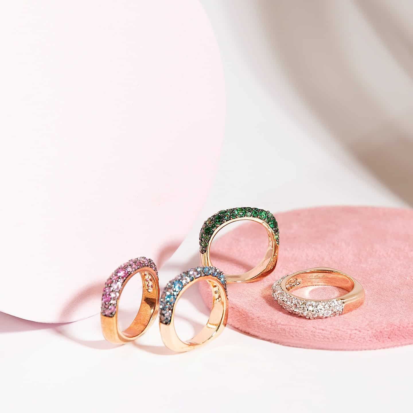 Quatre anneaux de pierres précieuses colorées sur des surfaces roses et blanches.