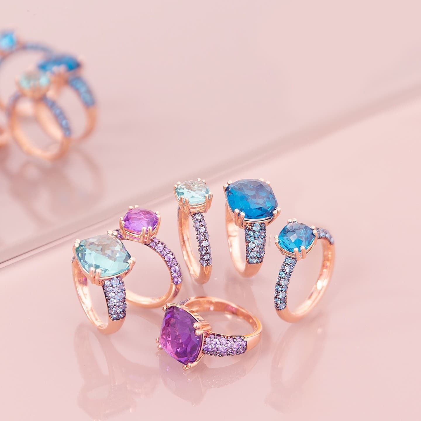 Six bagues en pierres précieuses colorées sur fond rose. Anneaux en or rose ornés de pierres de différentes nuances de bleu et de violet.