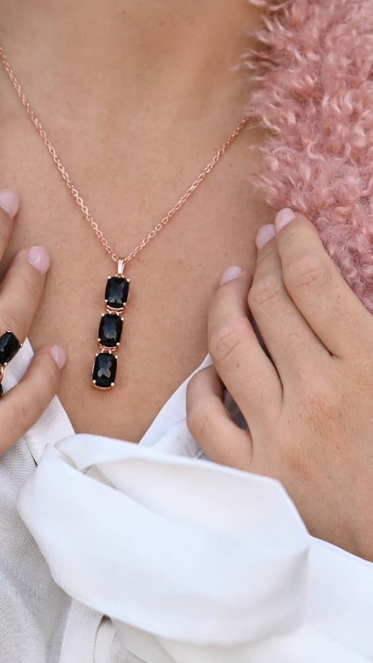 Femme portant un collier en or rose avec trois pierres rectangulaires noires, mains sur la poitrine, chemise blanche, fourrure rose.