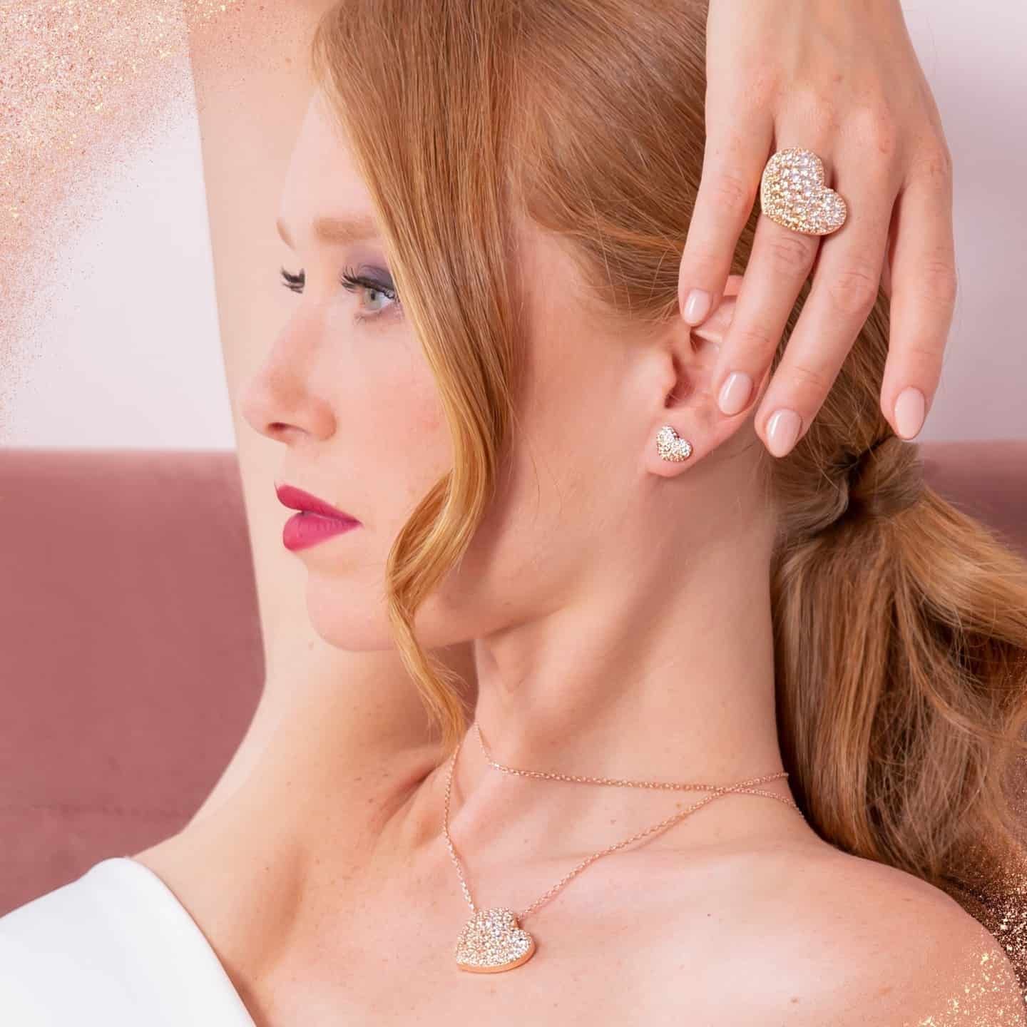 Femme aux cheveux roux portant des bijoux en forme de cœur, du rouge à lèvres rose, posant sur un fond rose.