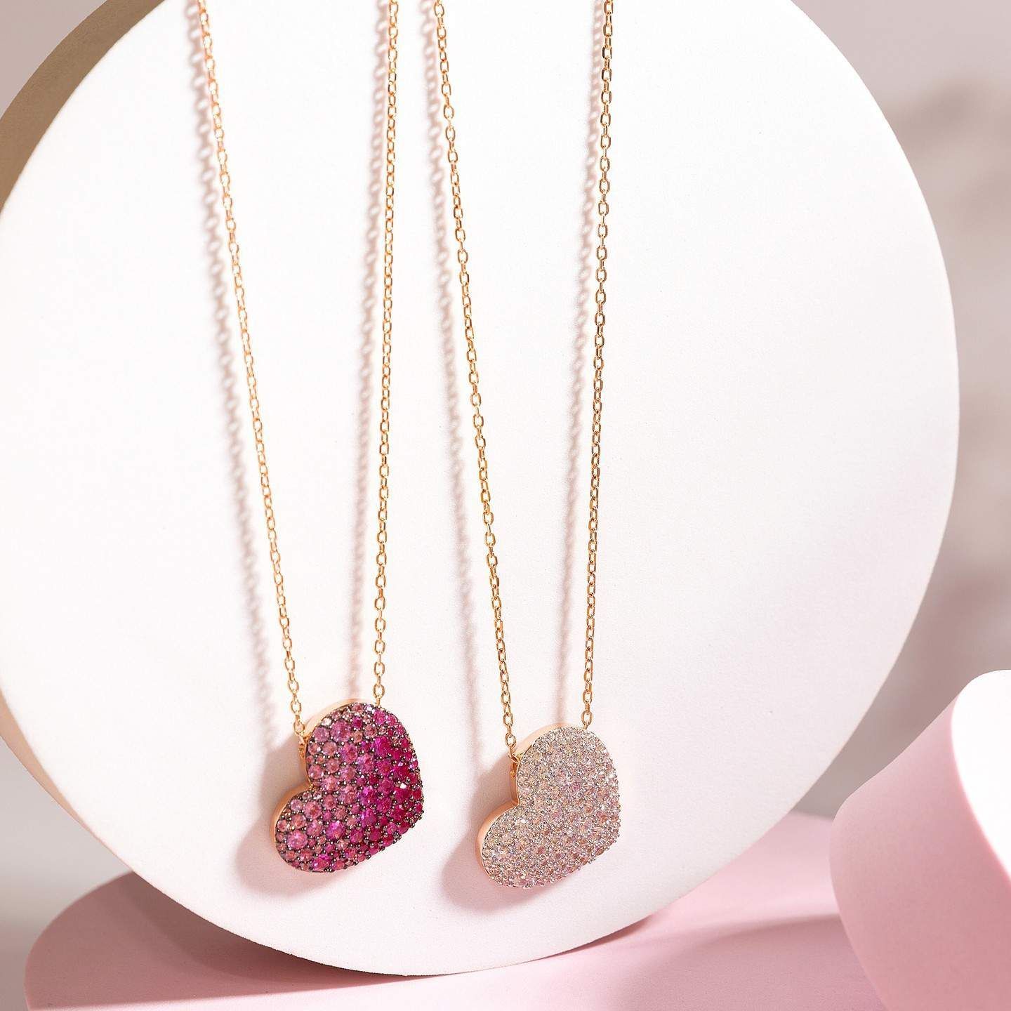 Deux colliers en or avec pendentifs en forme de cœur recouverts de pierres précieuses roses et blanches, sur fond blanc.