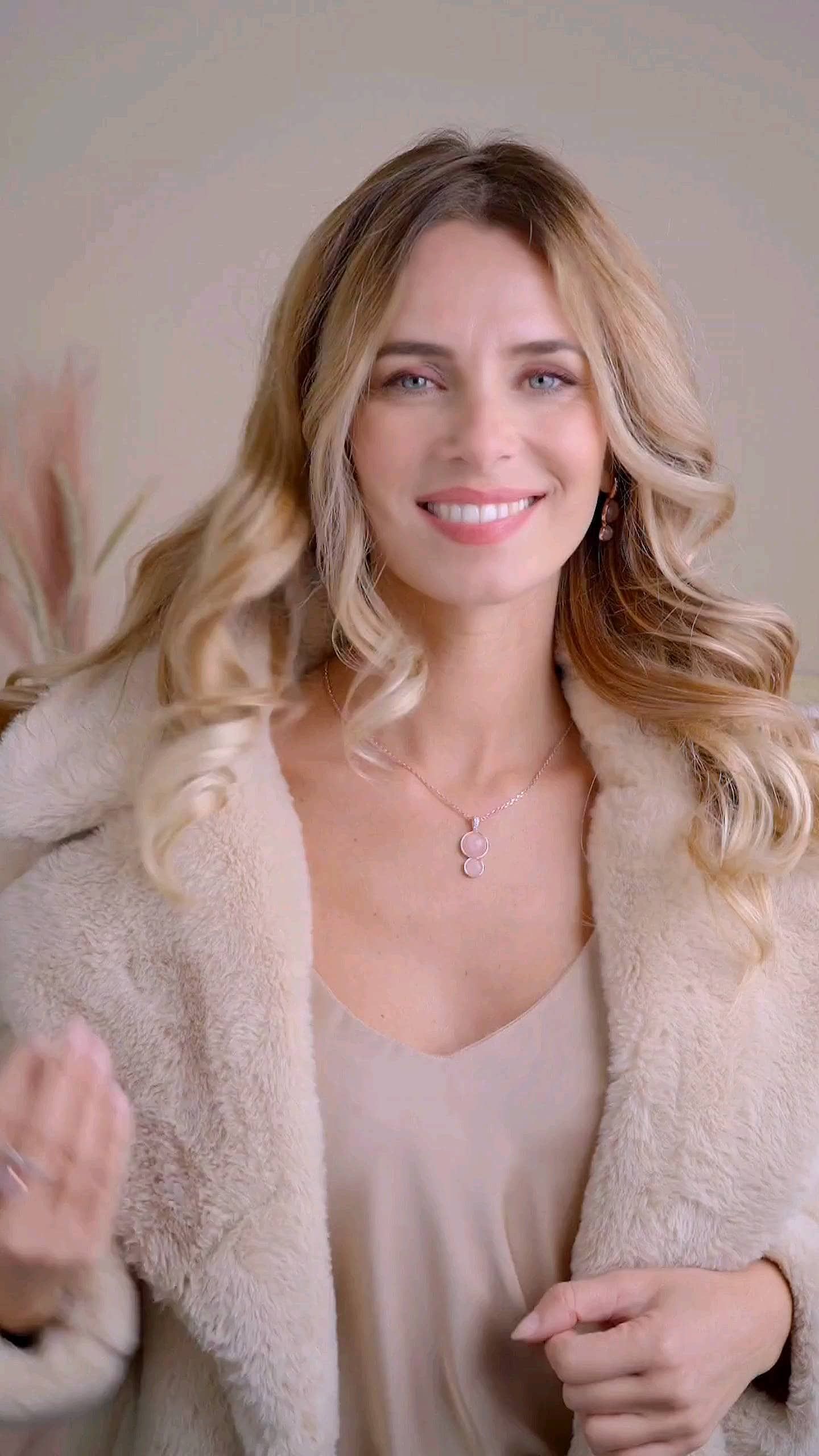 Femme blonde souriante, portant un manteau et un collier de couleur crème, à l'intérieur.