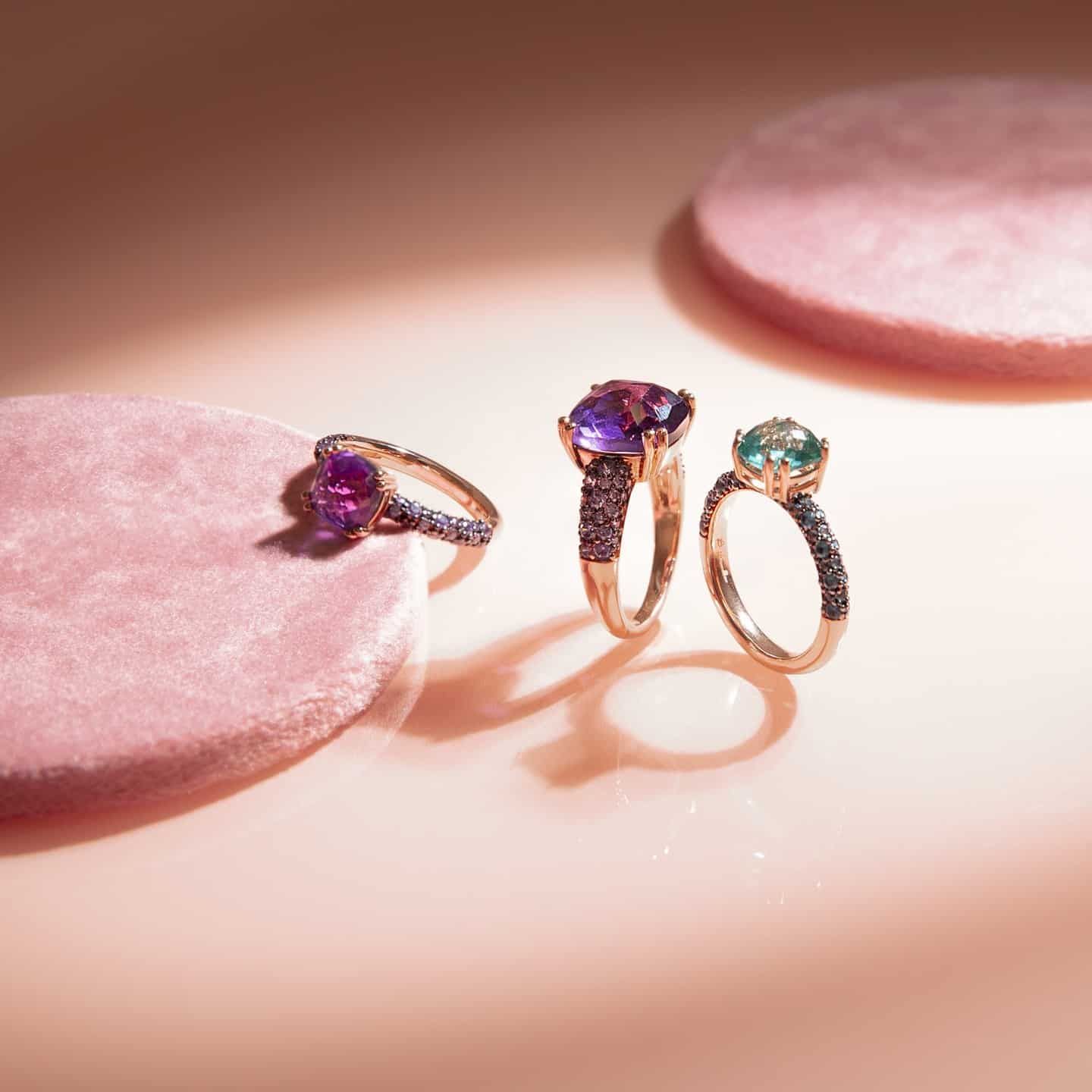 Trois bagues en pierres précieuses sur fond rose. Améthyste et pierres bleu-vert serties sur or.