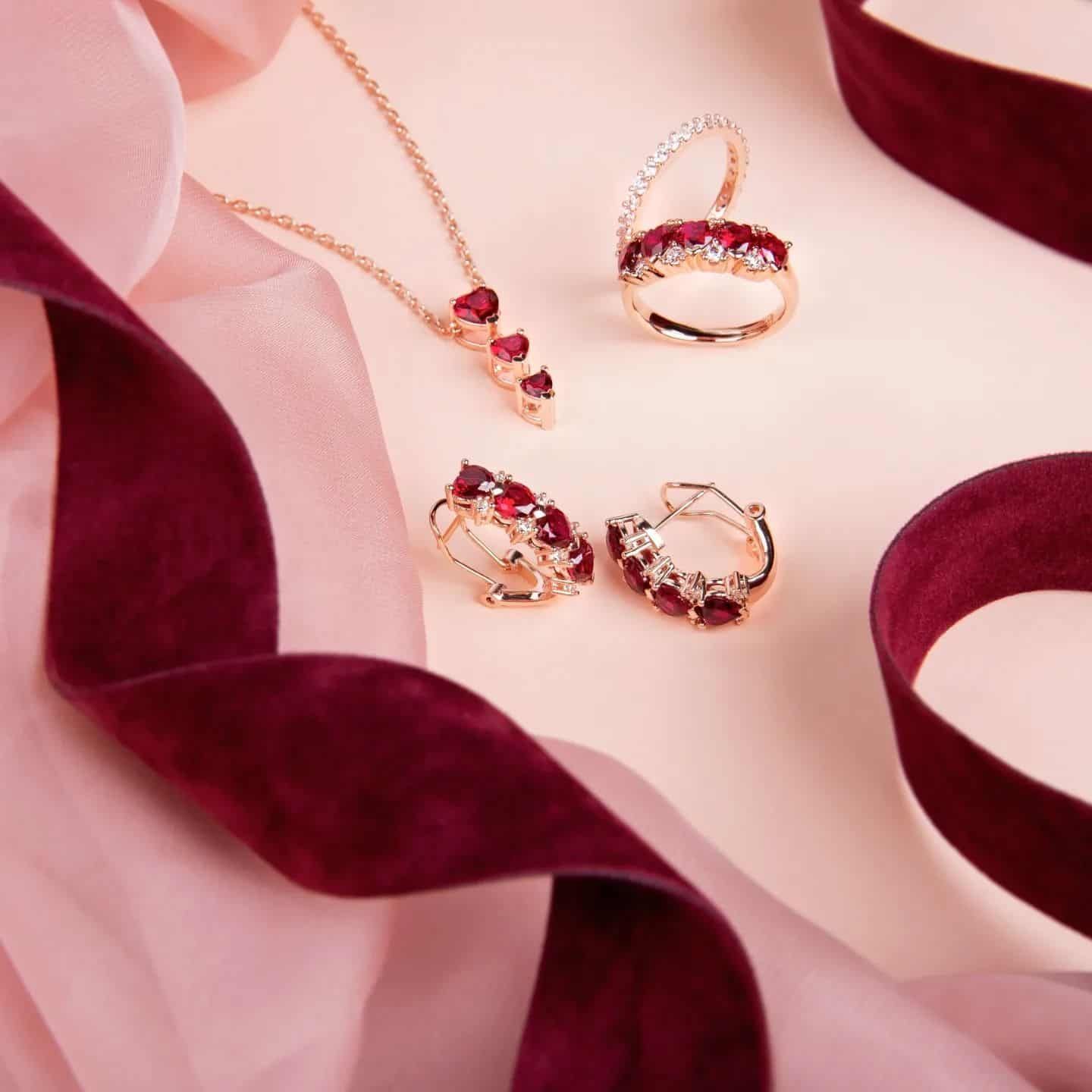 Parure de bijoux avec pierres rouges et or : collier, bagues et boucles d'oreilles, avec ruban bordeaux sur fond rose.
