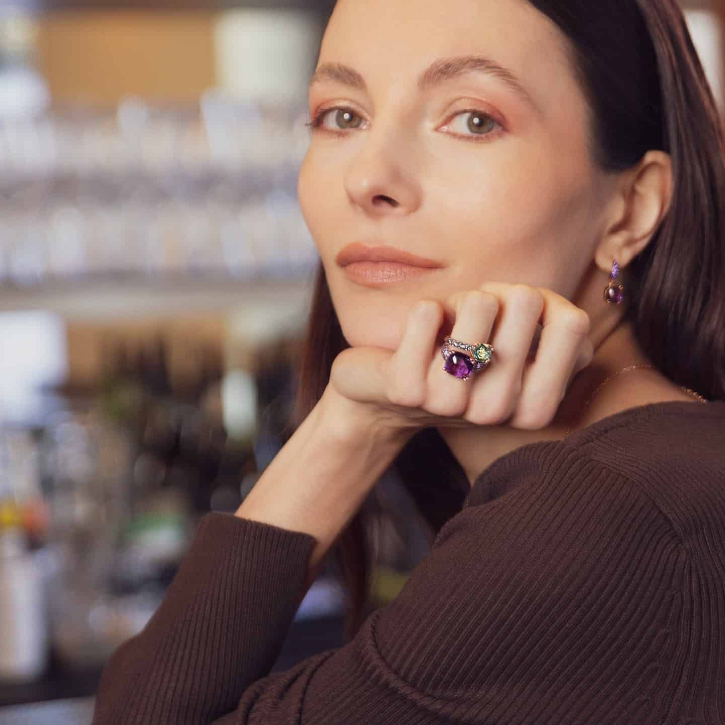 Femme avec bague et boucles d'oreilles en améthyste, haut marron, menton posé sur la main.