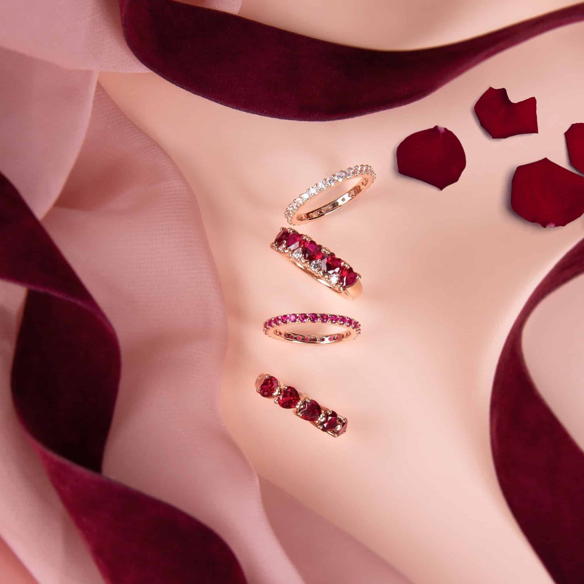 Quatre bagues en or avec diamants et rubis sur une surface crème avec ruban bordeaux et pétales de rose.