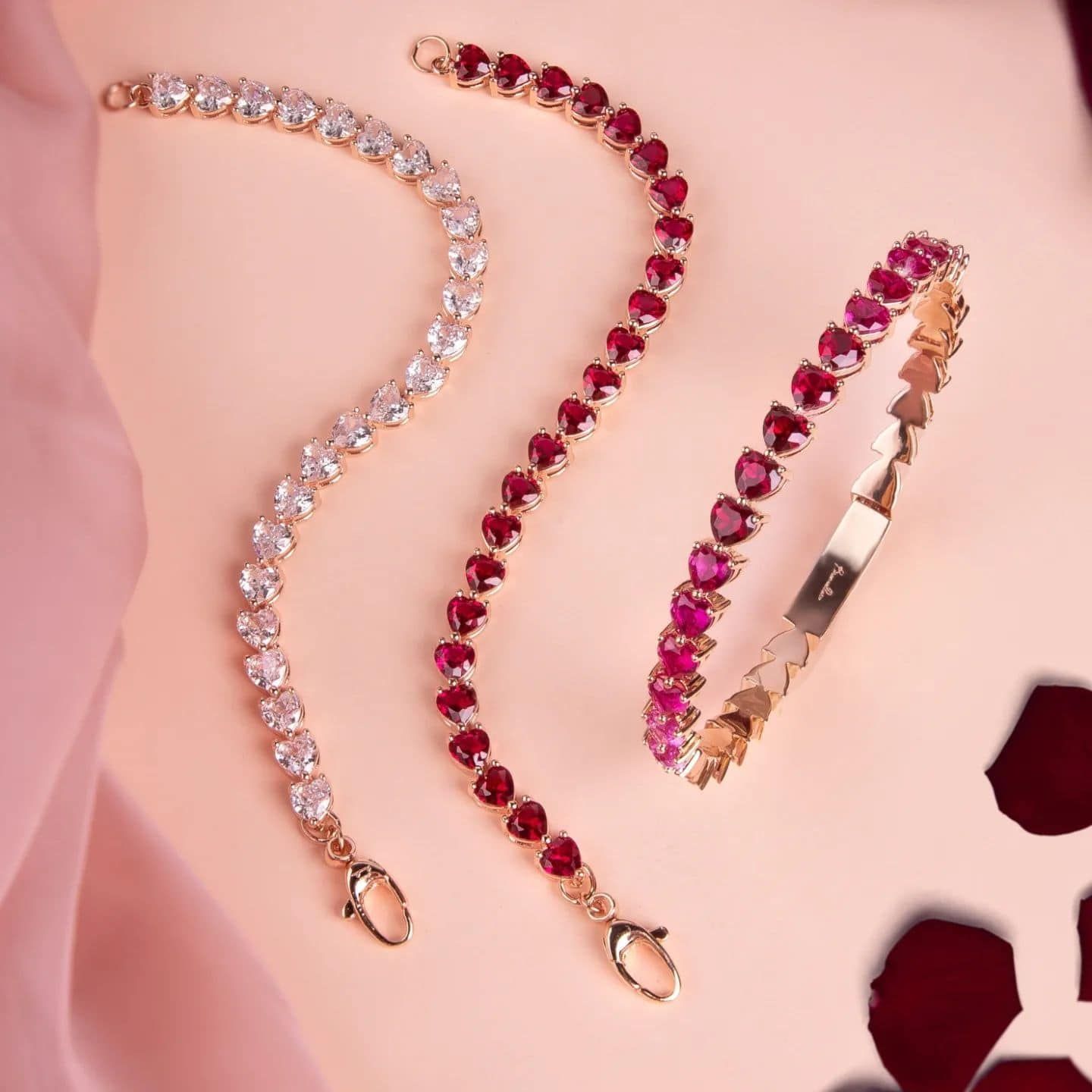 Trois bracelets ornés de pierres précieuses : un avec des pierres claires, deux avec des pierres précieuses rouges en forme de cœur, présentés sur une surface rose.