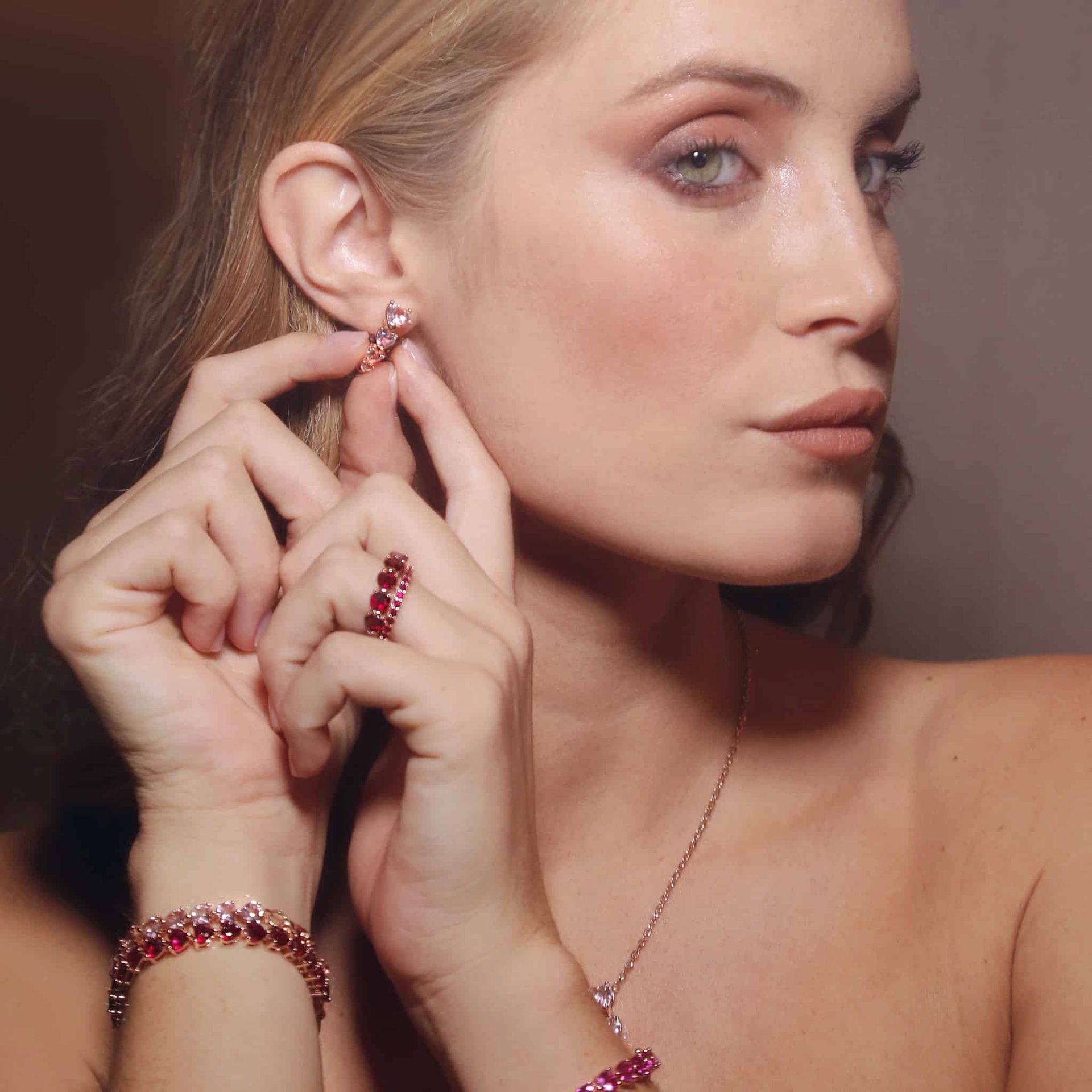 Femme aux cheveux blonds mettant des boucles d'oreilles, portant des bijoux en rubis : bracelet, bague et collier.