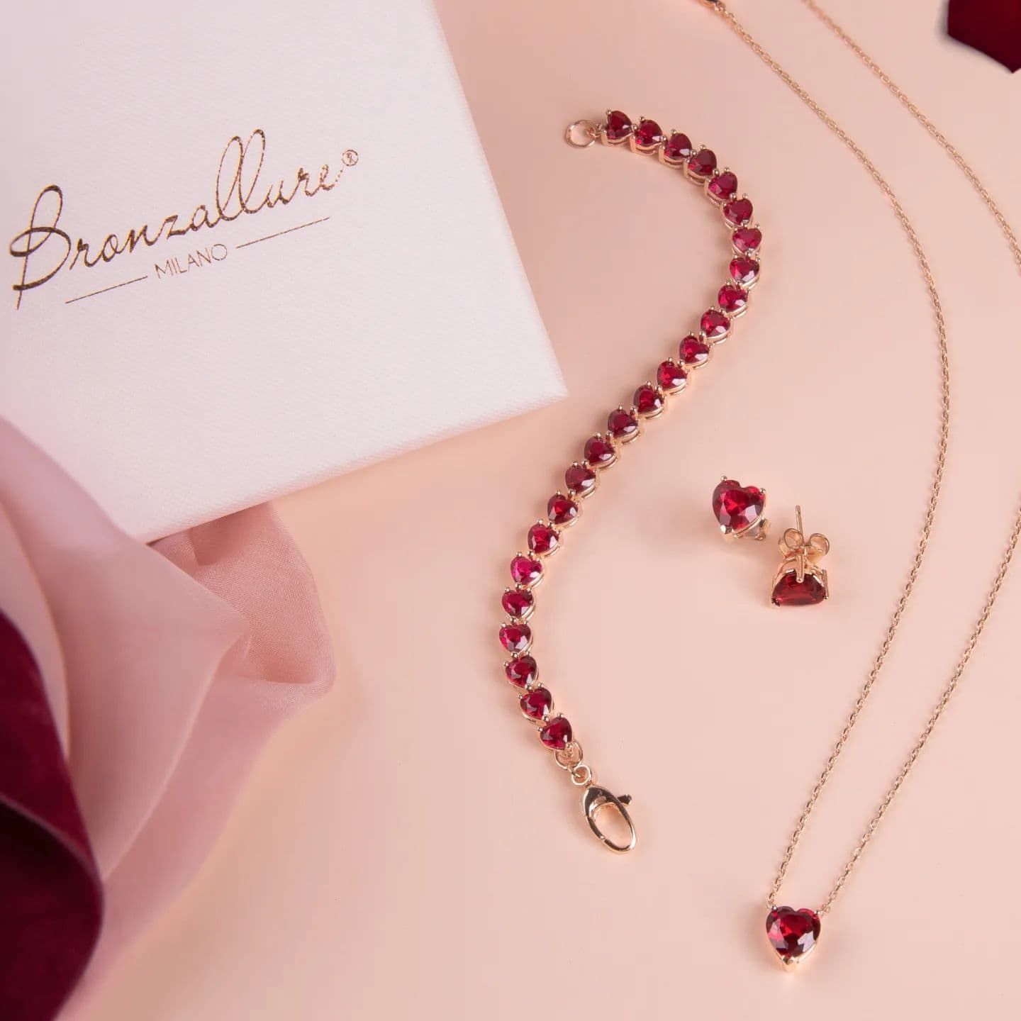 Parure de bijoux avec pierres précieuses rouges en forme de coeur et chaîne en or sur une surface rose, avec une boîte et un foulard.
