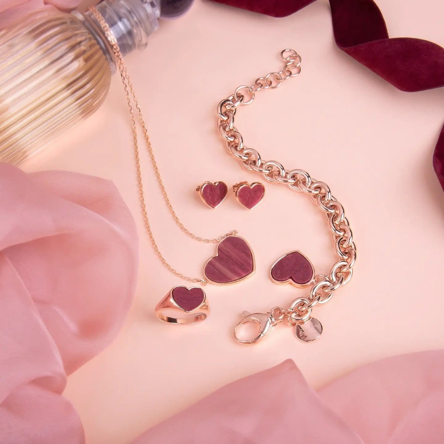 Parure de bijoux avec pendentifs bordeaux en forme de coeur : collier, bague, boucles d'oreilles et bracelet sur fond rose avec tissu et ruban.