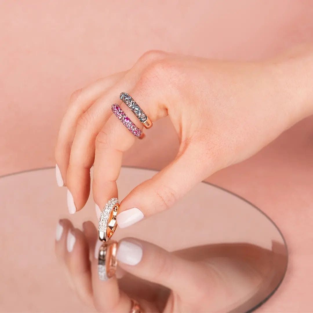 Main tenant trois bagues serties de pierres précieuses de différentes couleurs, au-dessus d'un miroir. Fond rose.