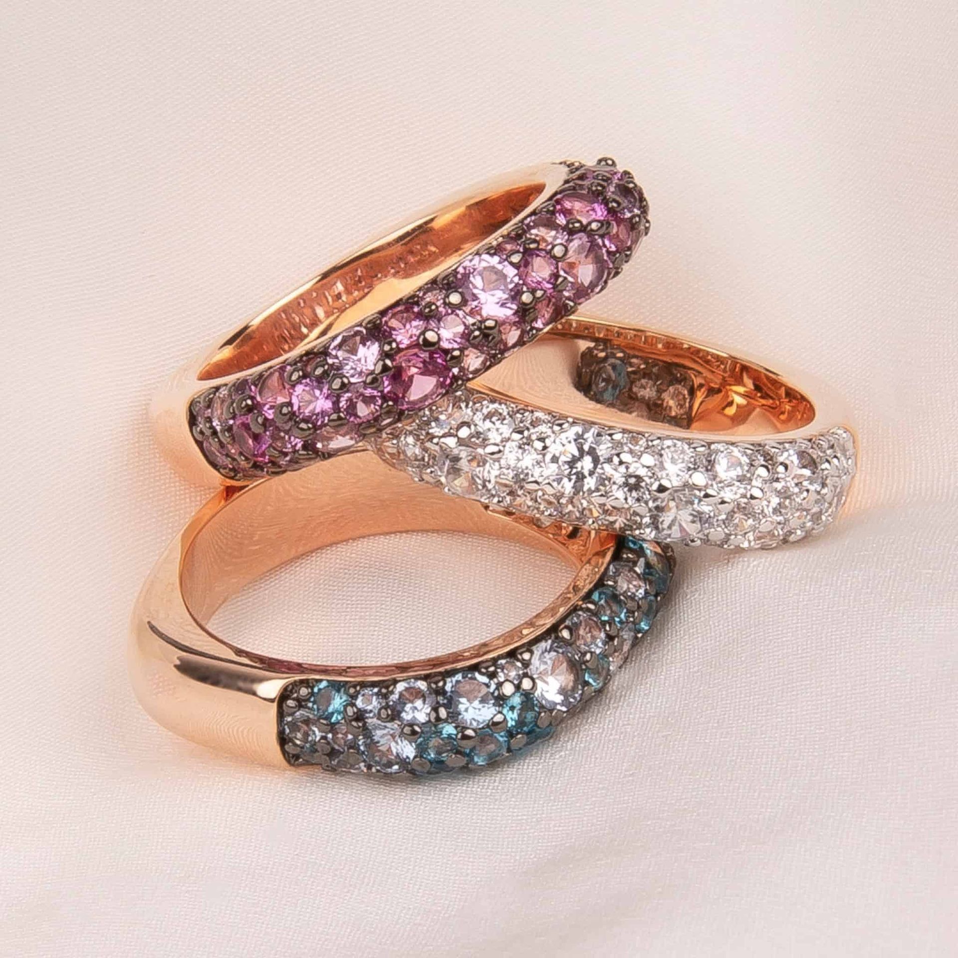 Trois bagues en or rose avec des pierres précieuses colorées, empilées sur un tissu blanc.