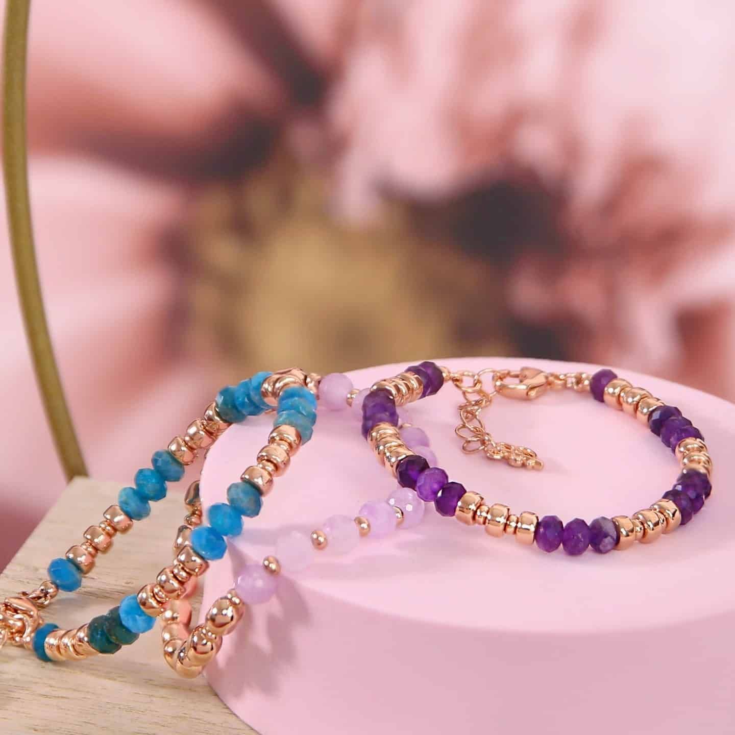 Trois bracelets de perles avec des accents en or rose sur un fond rose, mettant en valeur des perles bleues, violet clair et violet foncé.