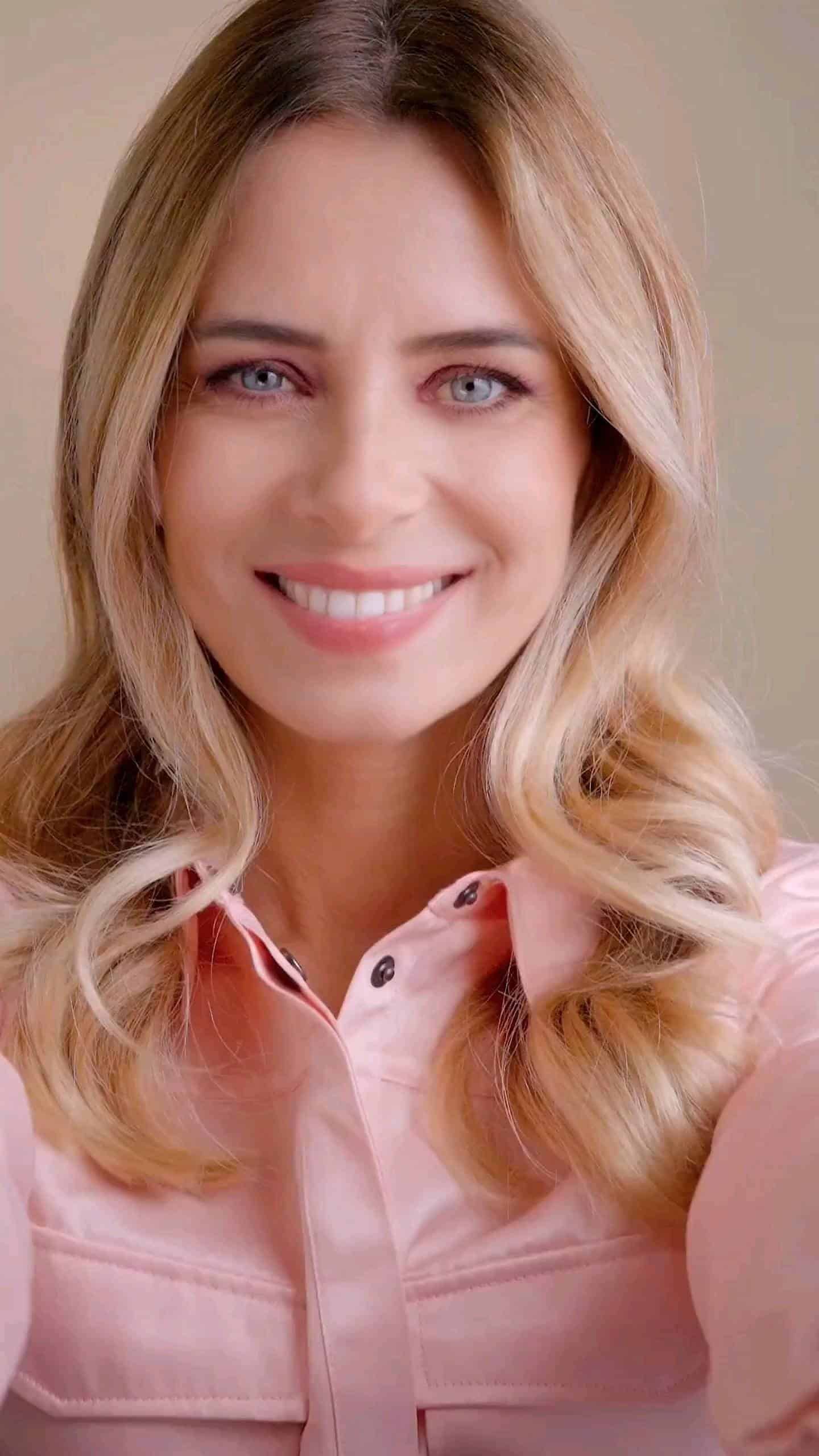 Femme aux cheveux blonds ondulés et aux yeux bleus, souriante dans une chemise boutonnée rose.