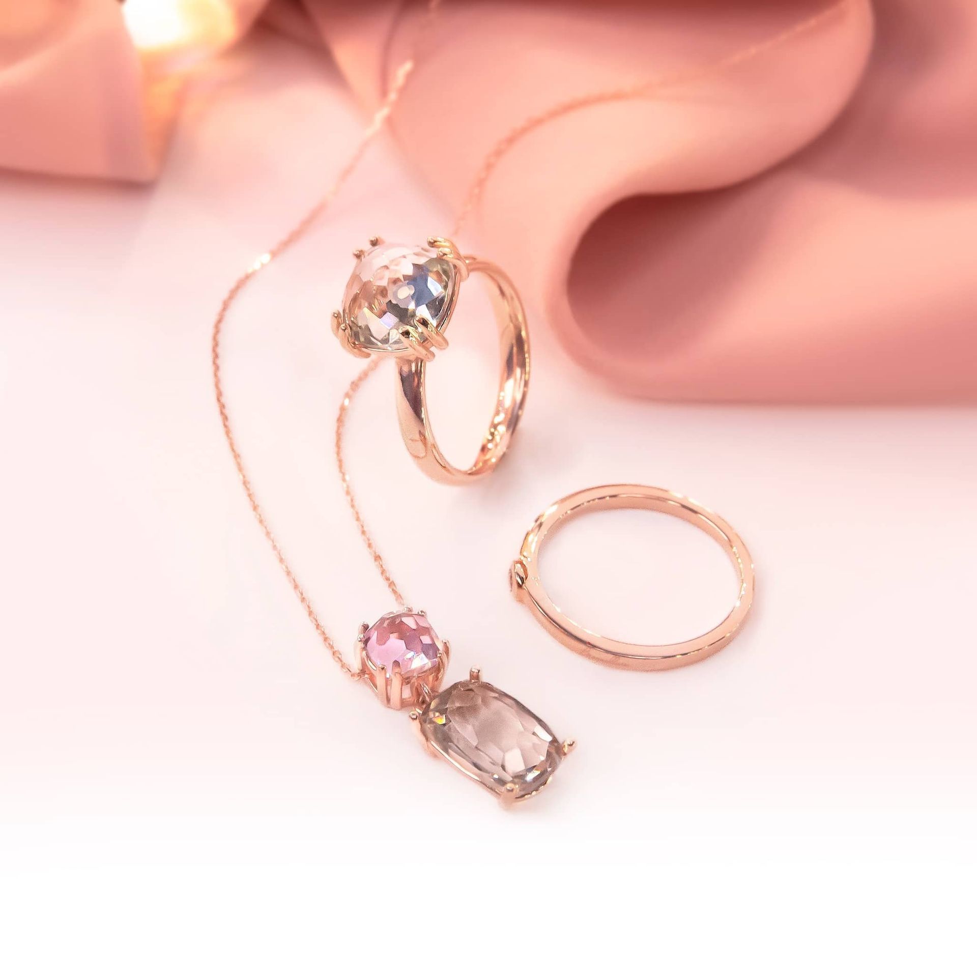 Ensemble de bijoux en or rose sur un tissu rose : une bague, un collier et une bague plus petite.