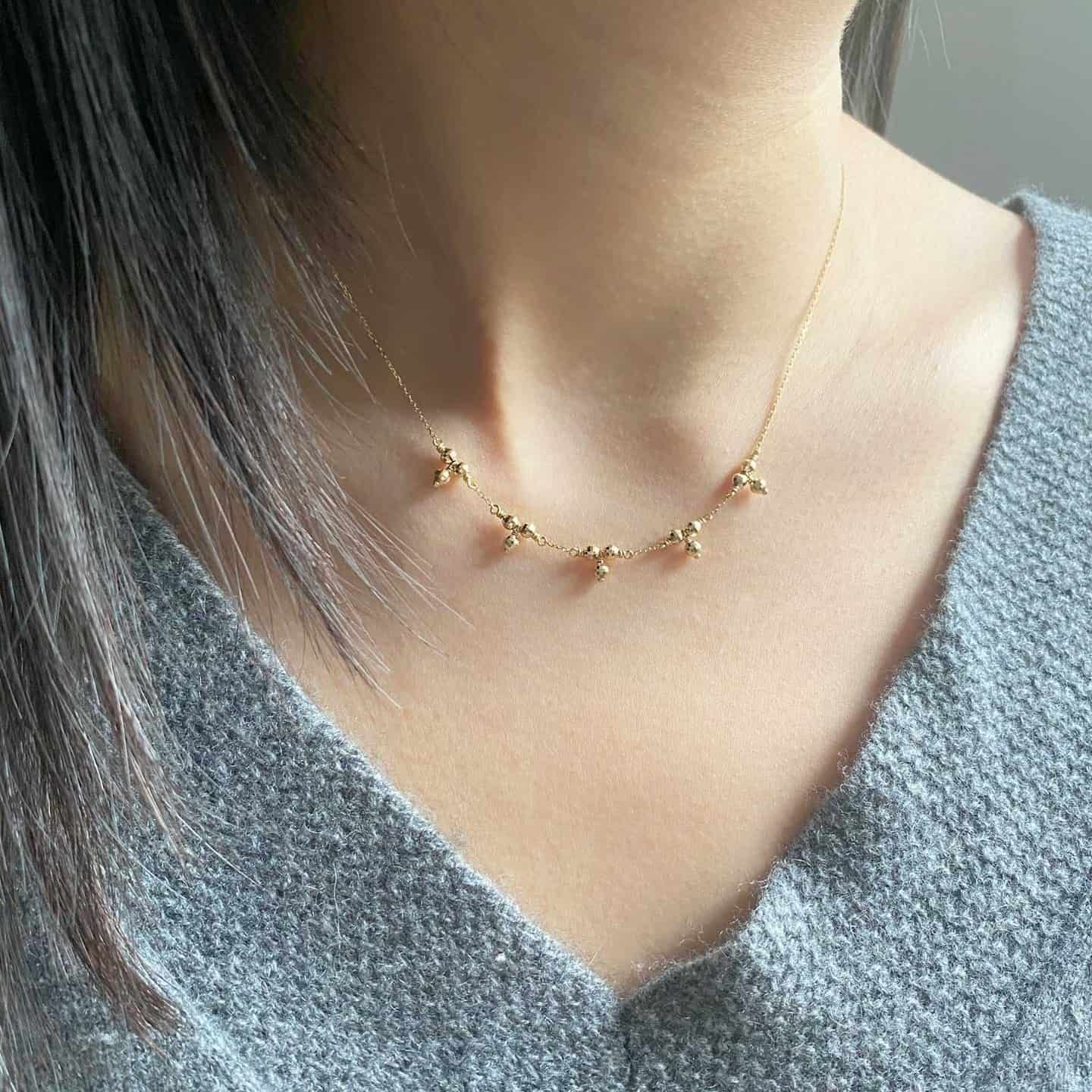 Femme portant un délicat collier en or avec de petites breloques en forme de boule, sur un pull gris.