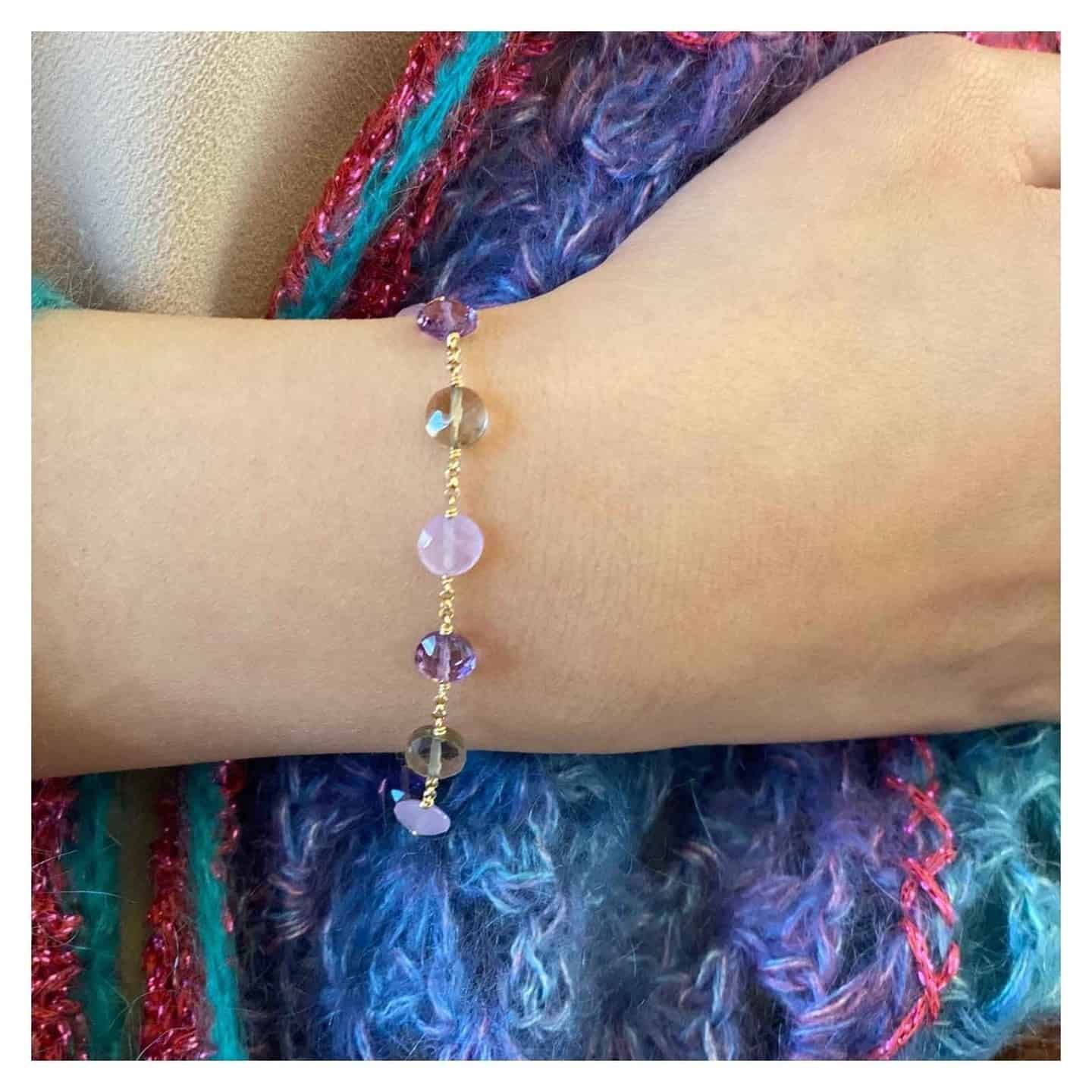 Un poignet orné d'un délicat bracelet en chaîne dorée orné de perles violettes et roses. Un foulard coloré en arrière-plan.