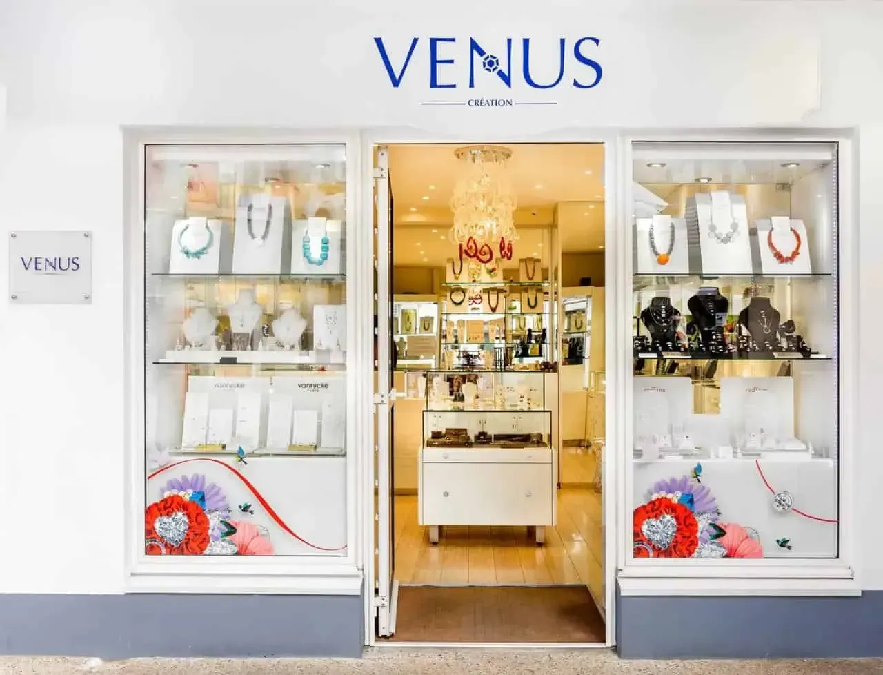 Entrée de la bijouterie : vitrines blanches avec bijoux, porte ouverte révélant d'autres présentoirs. Panneau bleu « VENUS » au-dessus.