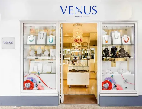Entrée de la bijouterie : vitrines blanches avec bijoux, porte ouverte révélant d'autres présentoirs. Panneau bleu « VENUS » au-dessus.