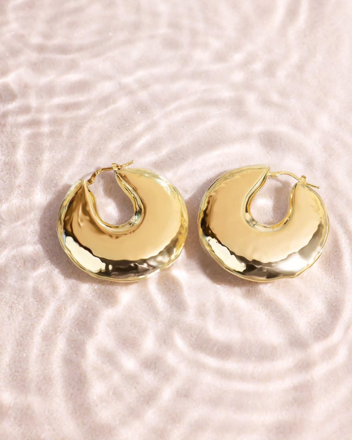 Boucles d'oreilles créoles rondes en or sur un fond rose clair avec des ondulations d'eau.