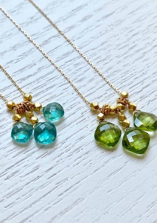 Deux délicats colliers en or, l'un avec des pierres précieuses bleues, l'autre avec des vertes, sur une surface texturée blanche.