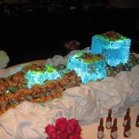 Blue lit ice sculptured cube decorations on a buffet table with shrimp and lemon wedges and red bottles nearby