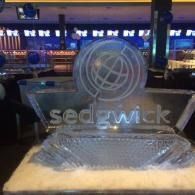 Ice sculpture with Sedgwick logo on a table in a dimly lit event venue.