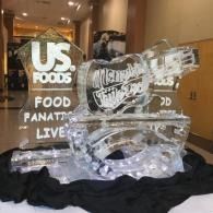 Clear ice sculpture on black cloth at a US Foods event display with illuminated signage in the background