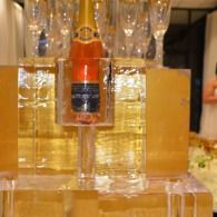 Bottle of orange drink in a clear dispenser, with stacked glasses behind it and a gold-toned backdrop.