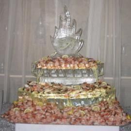 Ice sculptured tiered fruit display topped with a clear decorative sculpture, arranged on a table behind a curtain