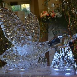 Clear ice bird sculpture with outstretched wings on a table, with flowers blurred in the background