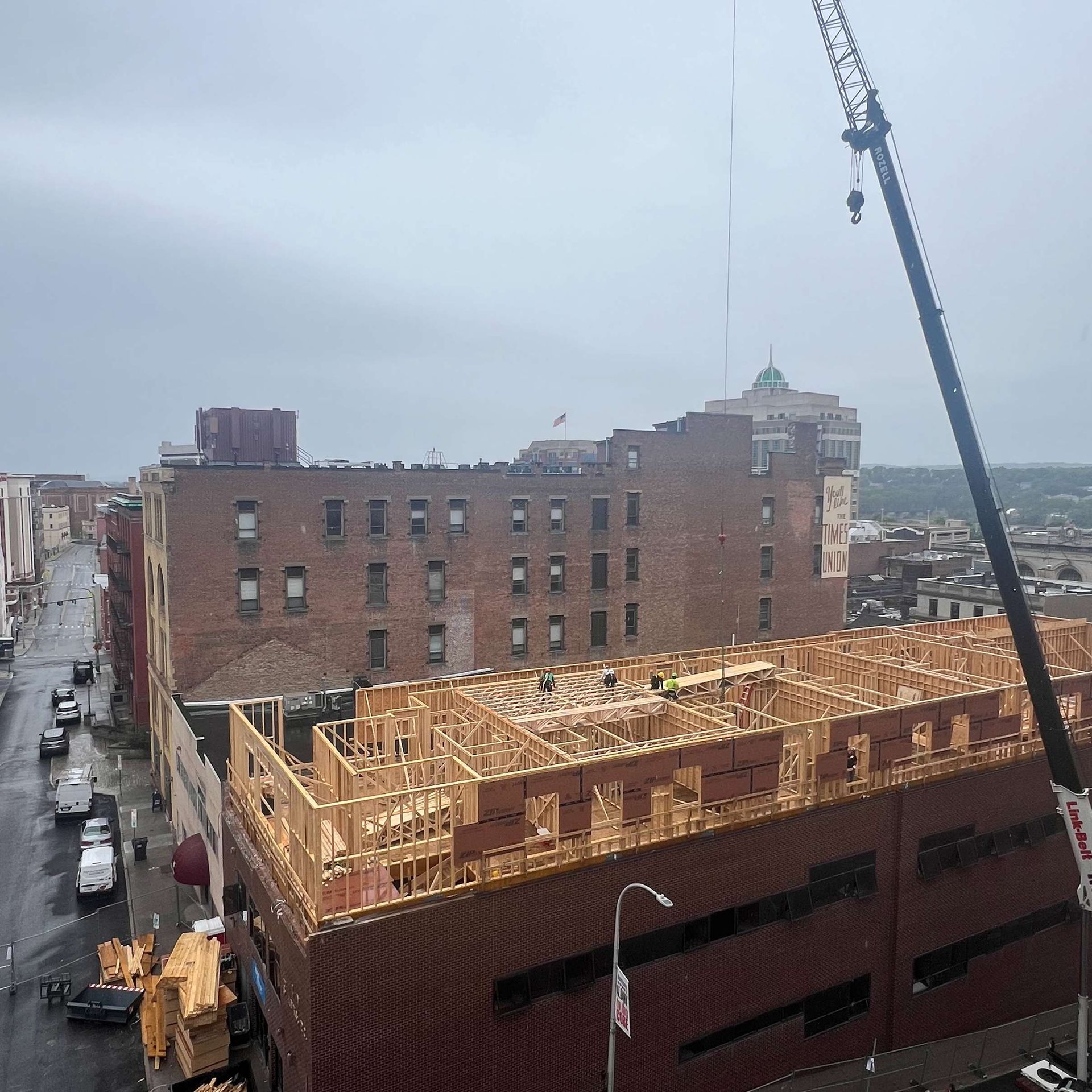 Construction site with wooden framing, brick buildings, and a crane. Workers are visible. Cloudy day.