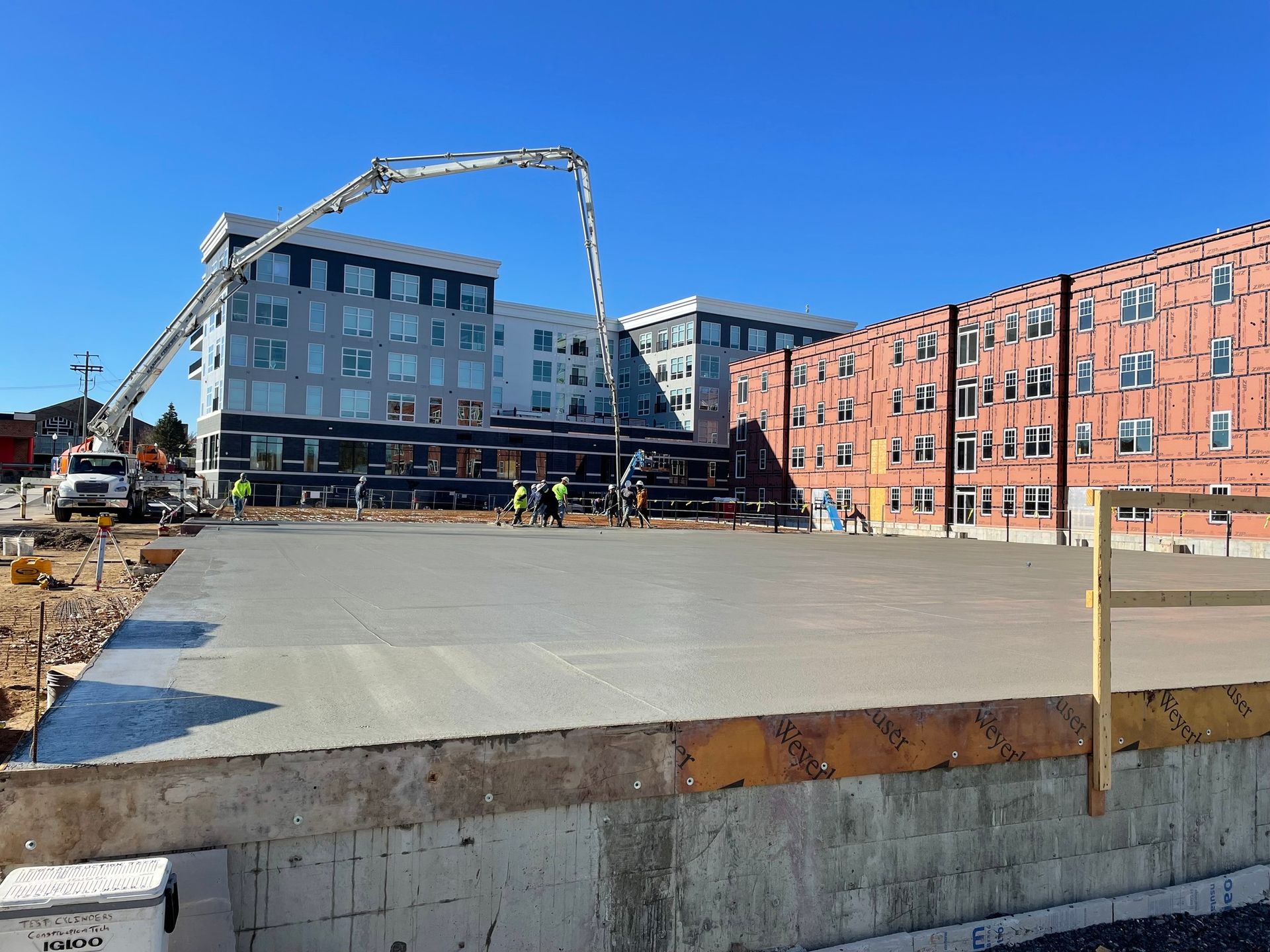 Concrete slab being poured at a construction site with buildings in the background and a concrete pump truck.