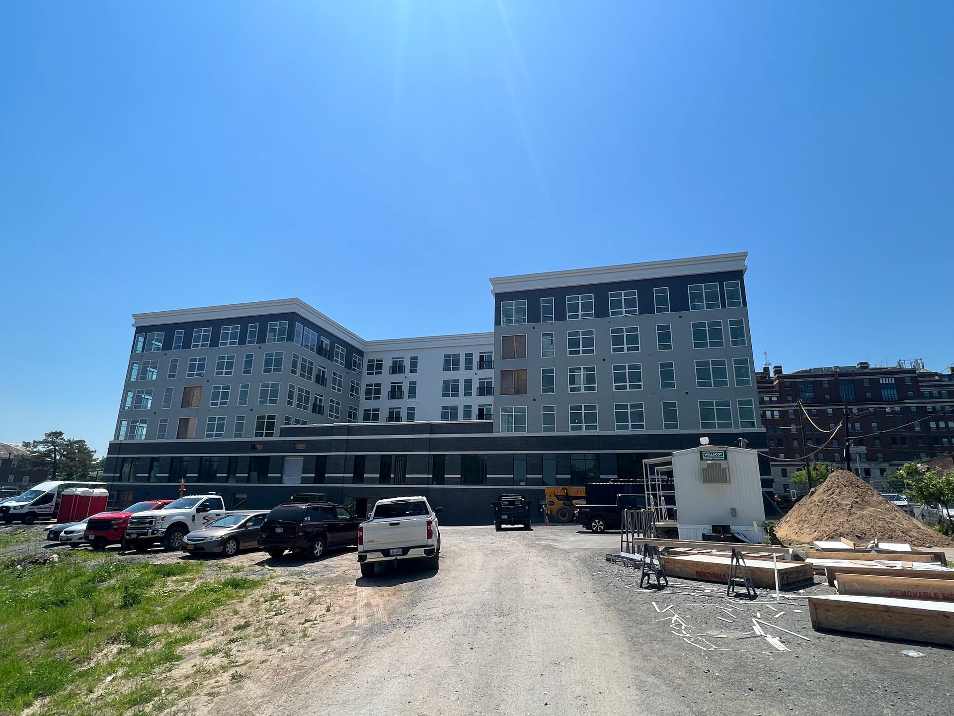 Multi-story apartment building with vehicles parked in front on a gravel lot under a bright blue sky.