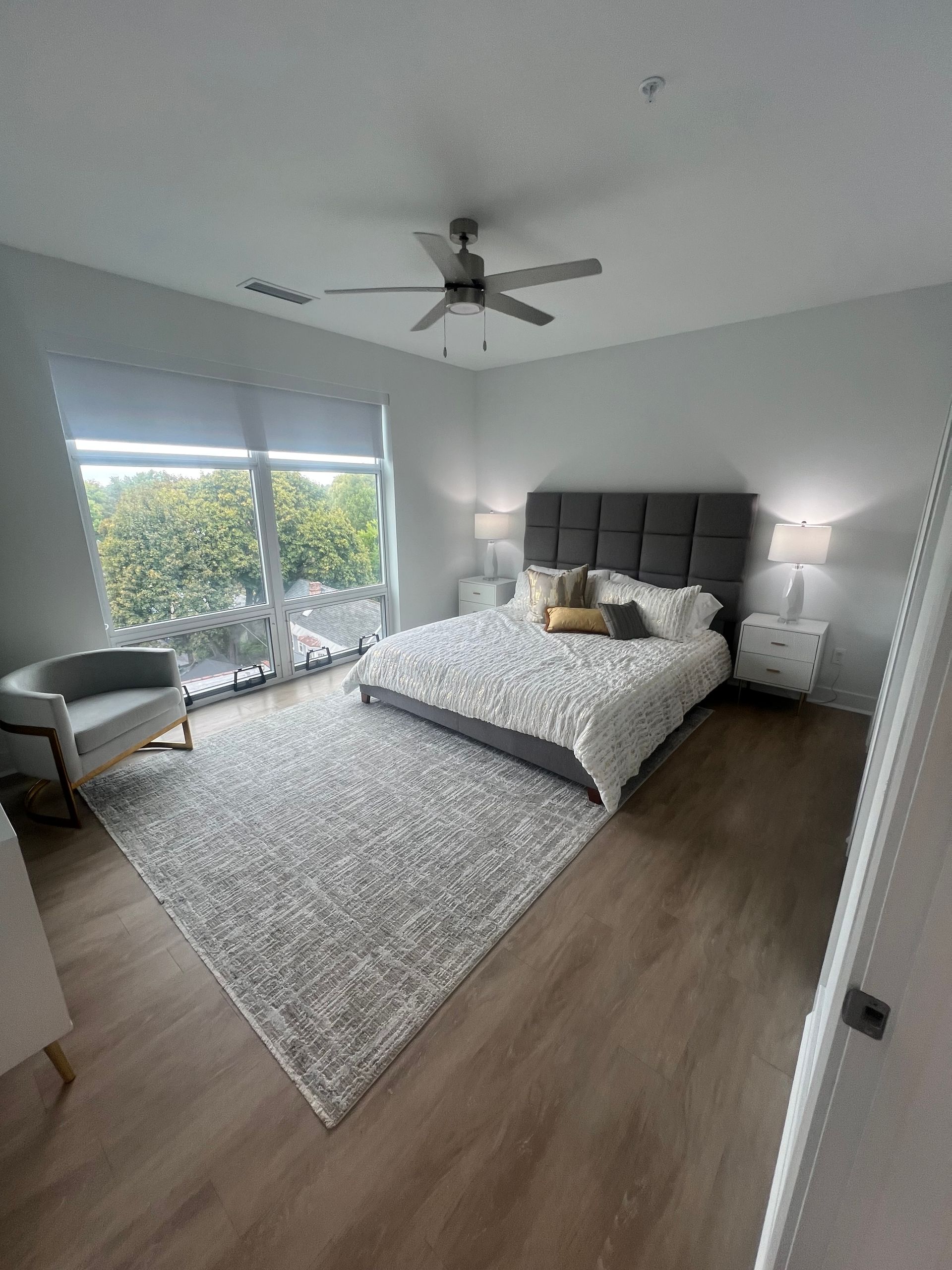 Bedroom with a gray bed, rug, chair, and white nightstands. Large window with shade. Ceiling fan.