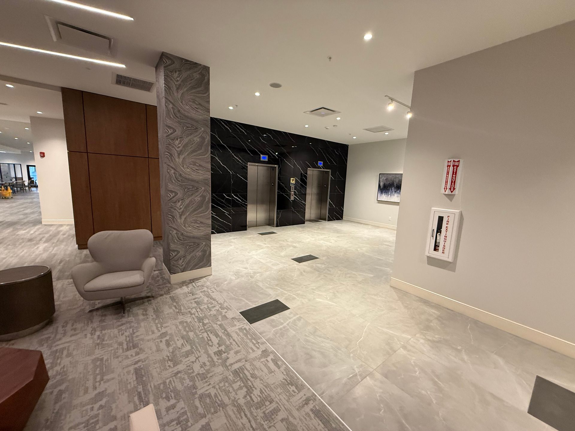 Lobby with elevators, seating area, and fire safety equipment. Gray and wood tones dominate the interior.