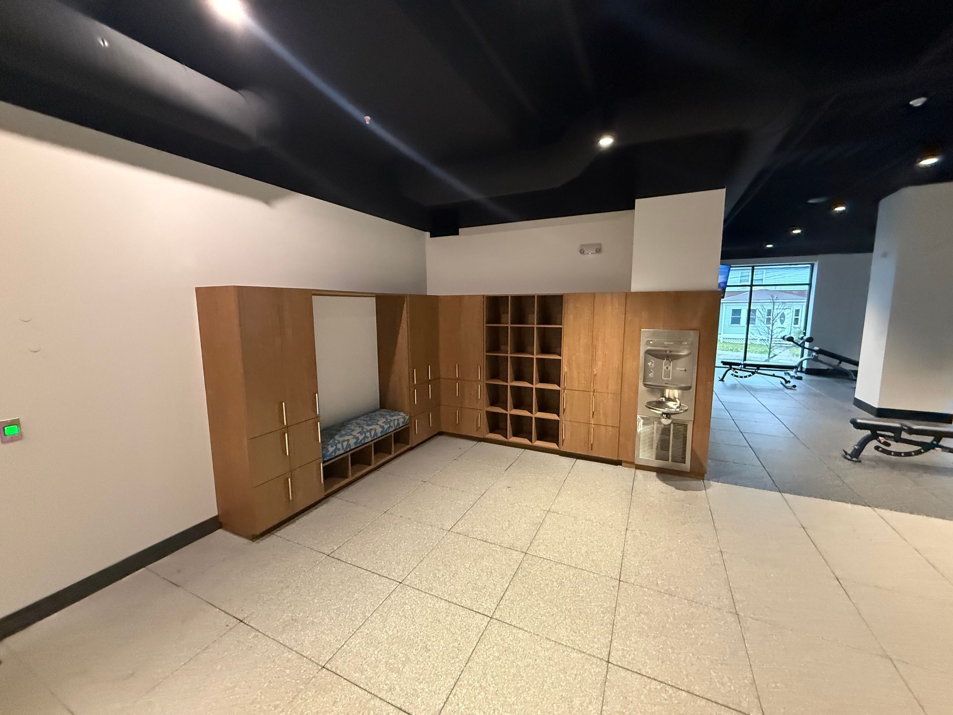 Locker room with wooden lockers, shoe cubbies, and a bench. Light-colored tiled floor, black ceiling, and natural light.
