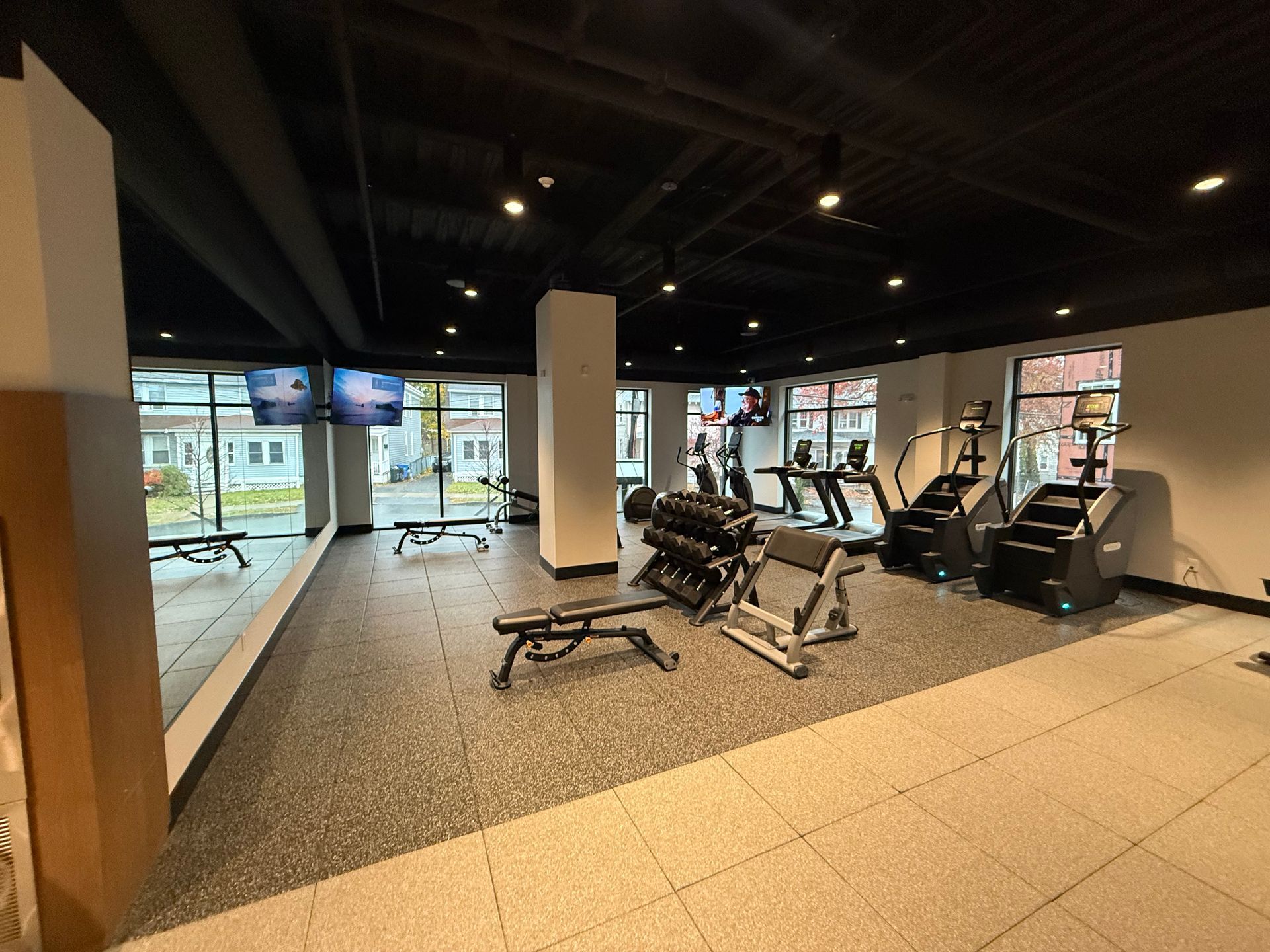 Gym interior with fitness equipment: treadmills, weights, and stair climbers. Large windows and black ceiling.