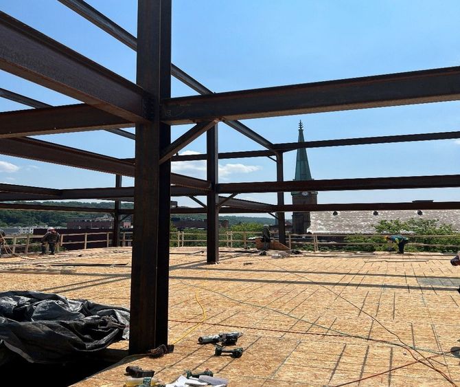 Steel beams frame a building under construction. Workers on plywood floor with church steeple visible in background.