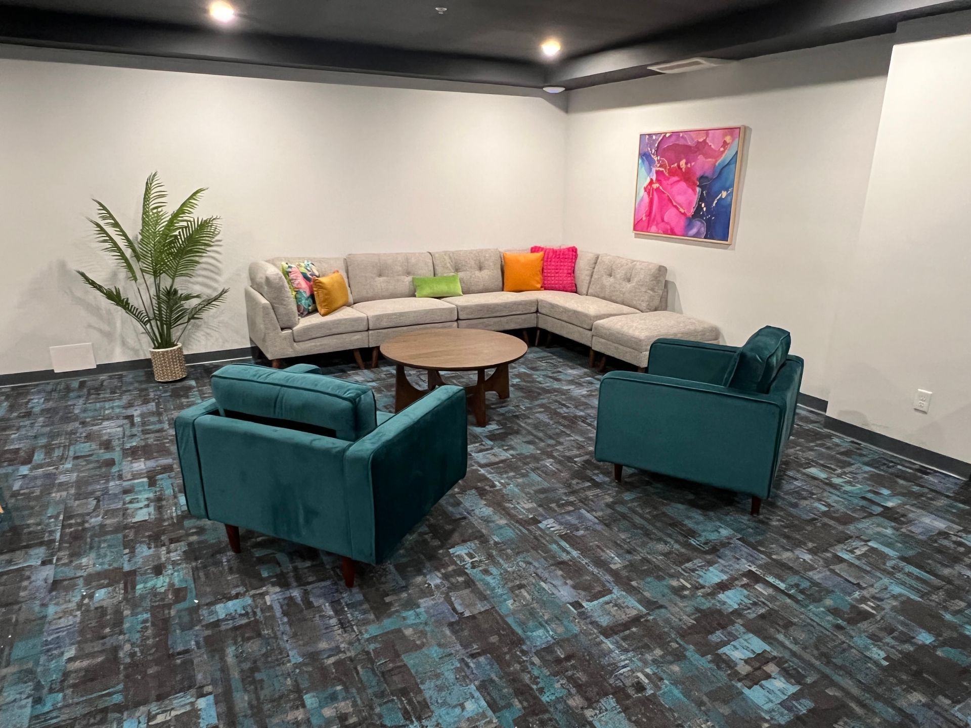 A lounge area with a sectional sofa, two teal armchairs, a coffee table, and a colorful abstract painting.