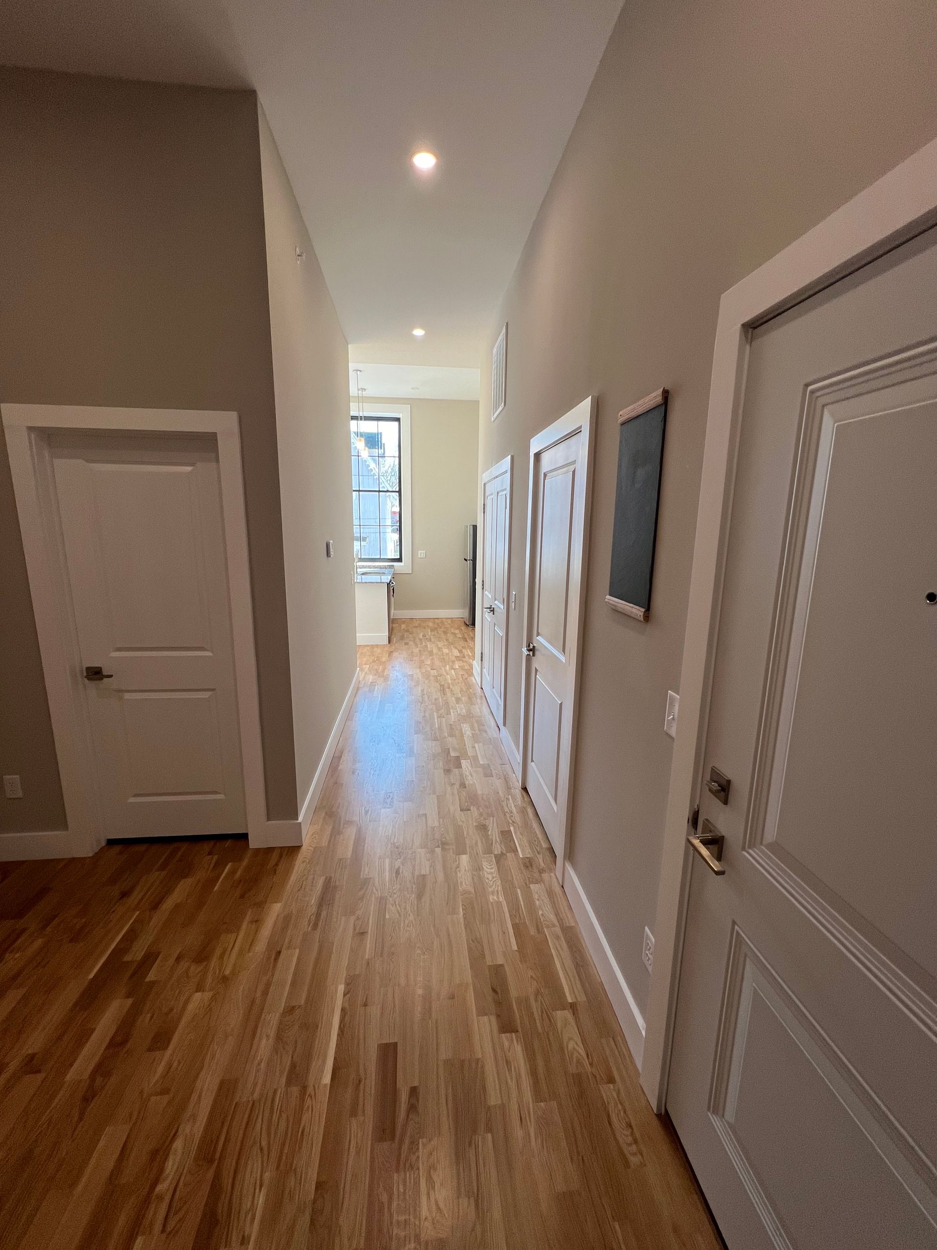 Long hallway with hardwood floors, white doors, and a distant window.