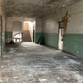 Interior of a dilapidated building with peeling paint, a staircase, and a light-filled doorway.